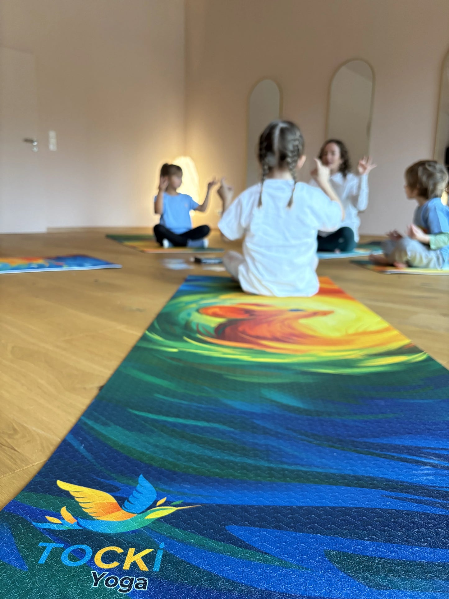 Bunte Kinderyogamatte mit Maus-Motiv, im Hintergrund Kinder im Kreis bei Yogaübung sitzend